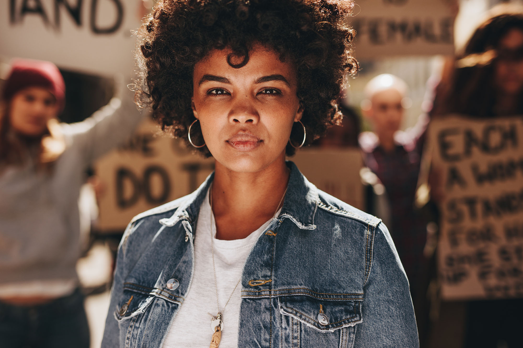 Young Black woman standing outdoors with group of demonstrator in background.