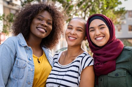 Group of three happy multiethnic friends looking at camera. Portrait of young women of different cultures enjoying vacation together. Smiling islamic girl with two african american friends outdoor.