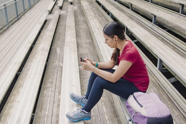 A student sits on bleachers looking at a phone with a back pack to the side